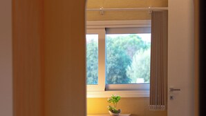 Ein Zimmer mit einem Bett, einem Vorhangfenster, einem kleinen Tisch mit Pflanze und einem Spiegel, der den Raum reflektiert.