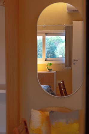 Ein Zimmer mit einem Bett, einem Vorhangfenster, einem kleinen Tisch mit Pflanze und einem Spiegel, der den Raum reflektiert.