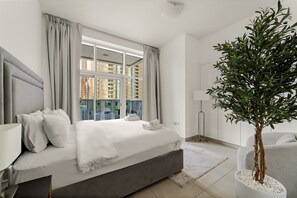 A modern bedroom with a large bed, a potted plant, and a view of buildings through the window. - Marina Arcade - 702 2BD (Dubai)