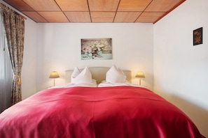 A bed with a red cover, two bedside lamps, and a floral painting on the wall. - Hotel Bären - Three-Star: Double Room 2 / Breakfast Included / 2 Guests / WiFi (Bernau im Schwarzwald)
