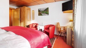 A hotel room with a bed, a red chair, a small table with a lamp, a TV mounted on the wall, and a painting of a mountain landscape. - Hotel Bären - Three-Star: Double Room 2 / Breakfast Included / 2 Guests / WiFi (Bernau im Schwarzwald)