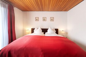 Habitación de hotel con techo de madera, una cama grande con ropa de cama roja, dos lámparas de noche y tres cuadros en la pared.