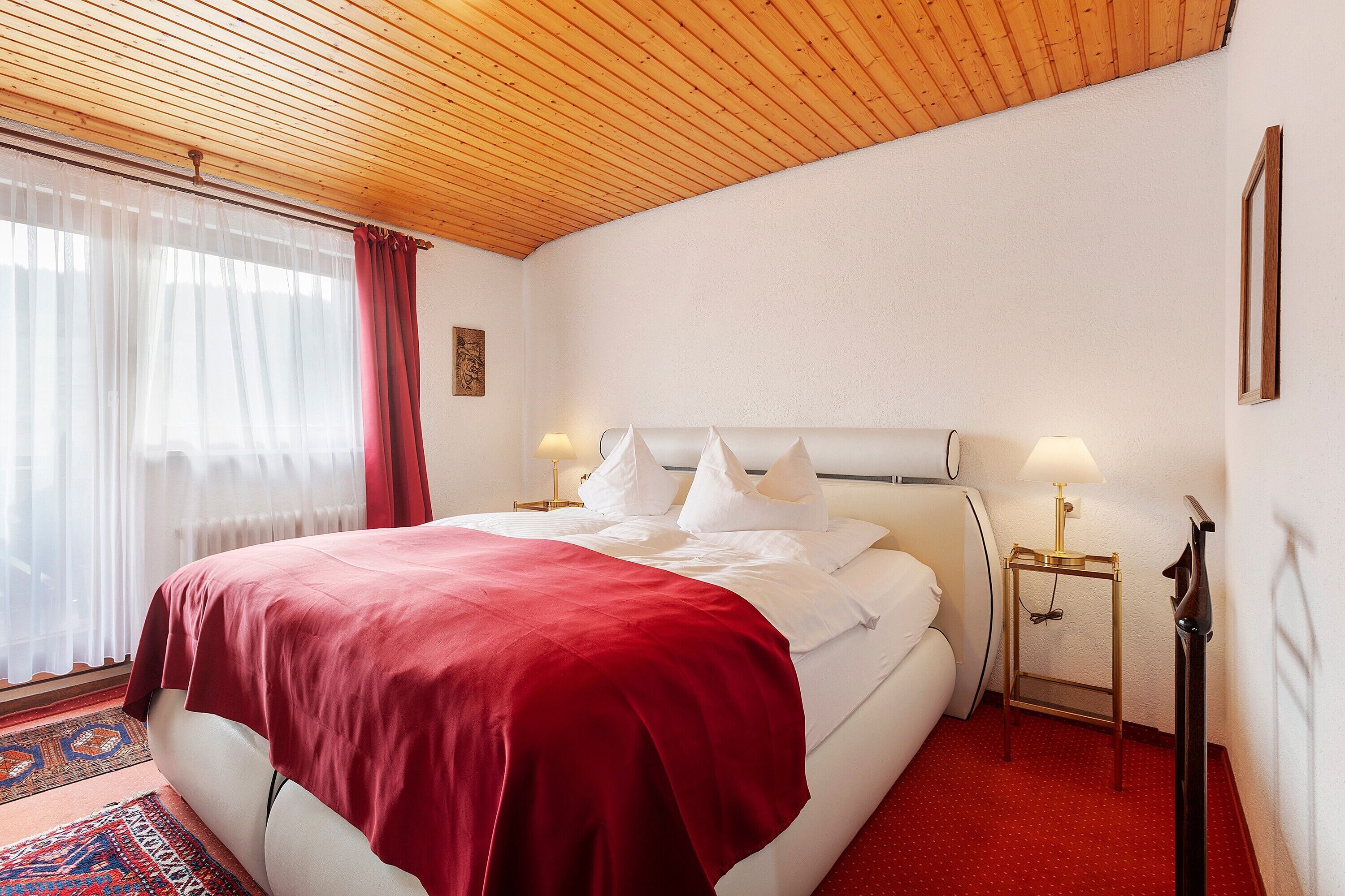 Hotellihuone, jossa on puinen katto, valkoisilla lakanoiden ja punaisella peitolla varustettu sänky, kaksi yöpöytävalaisinta ja punainen matto.