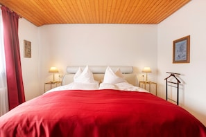 A hotel room with a wooden ceiling, a large bed with a red bedspread, two bedside tables with lamps, and a framed picture on the wall.