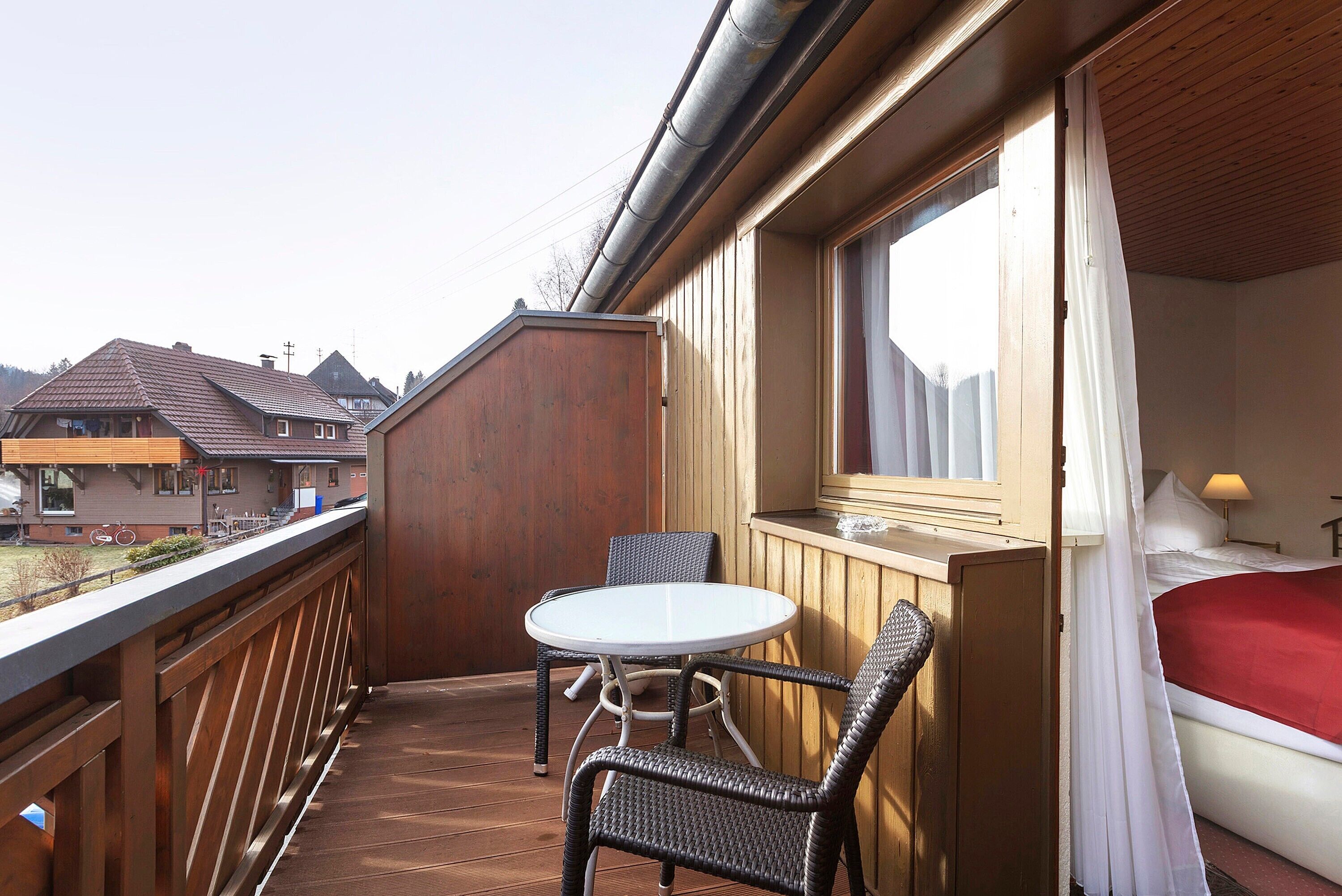 Un balcon avec une table et des chaises, un lit avec une couverture rouge et une fenêtre avec des rideaux.