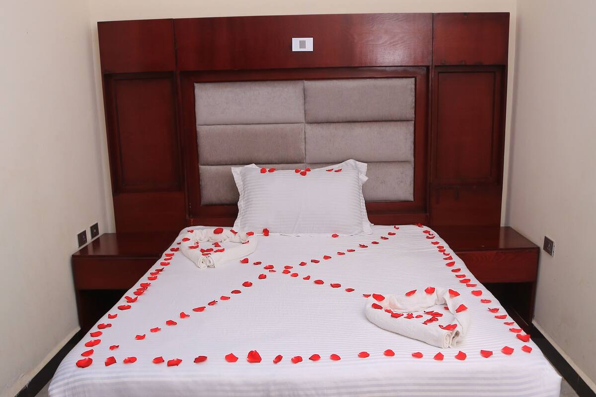 A neatly made bed with rose petals arranged on the bedspread, a wooden headboard, and a small rug on the floor.