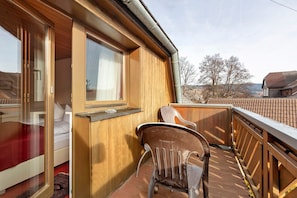 A wooden balcony with a chair, a bed with a red cover, and a window with curtains. - Hotel Bären - Three-Star Double Room 7 / incl. Breakfast / 3 Guests / Balcony with Mountain View (Bernau im Schwarzwald)