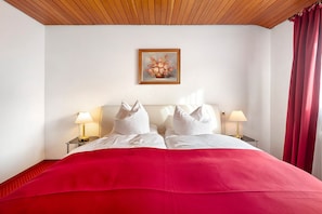 A hotel room with a wooden ceiling, white walls, a red bedspread, two white pillows, two bedside lamps, and a framed floral painting on the wall. - Hotel Bären - Three-Star Double Room 7 / incl. Breakfast / 3 Guests / Balcony with Mountain View (Bernau im Schwarzwald)