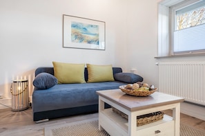 Ein Wohnzimmer mit einer Couch, Kissen, einem Tisch mit einem Korb, einem gerahmten Bild an der Wand und einem Fenster mit Jalousien.