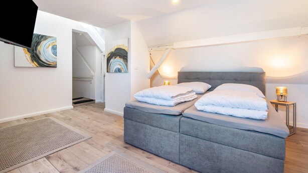 Une chambre à coucher avec un lit recouvert d’un tissu gris, une petite table de chevet avec une lampe et des œuvres d’art abstraites accrochées au mur.