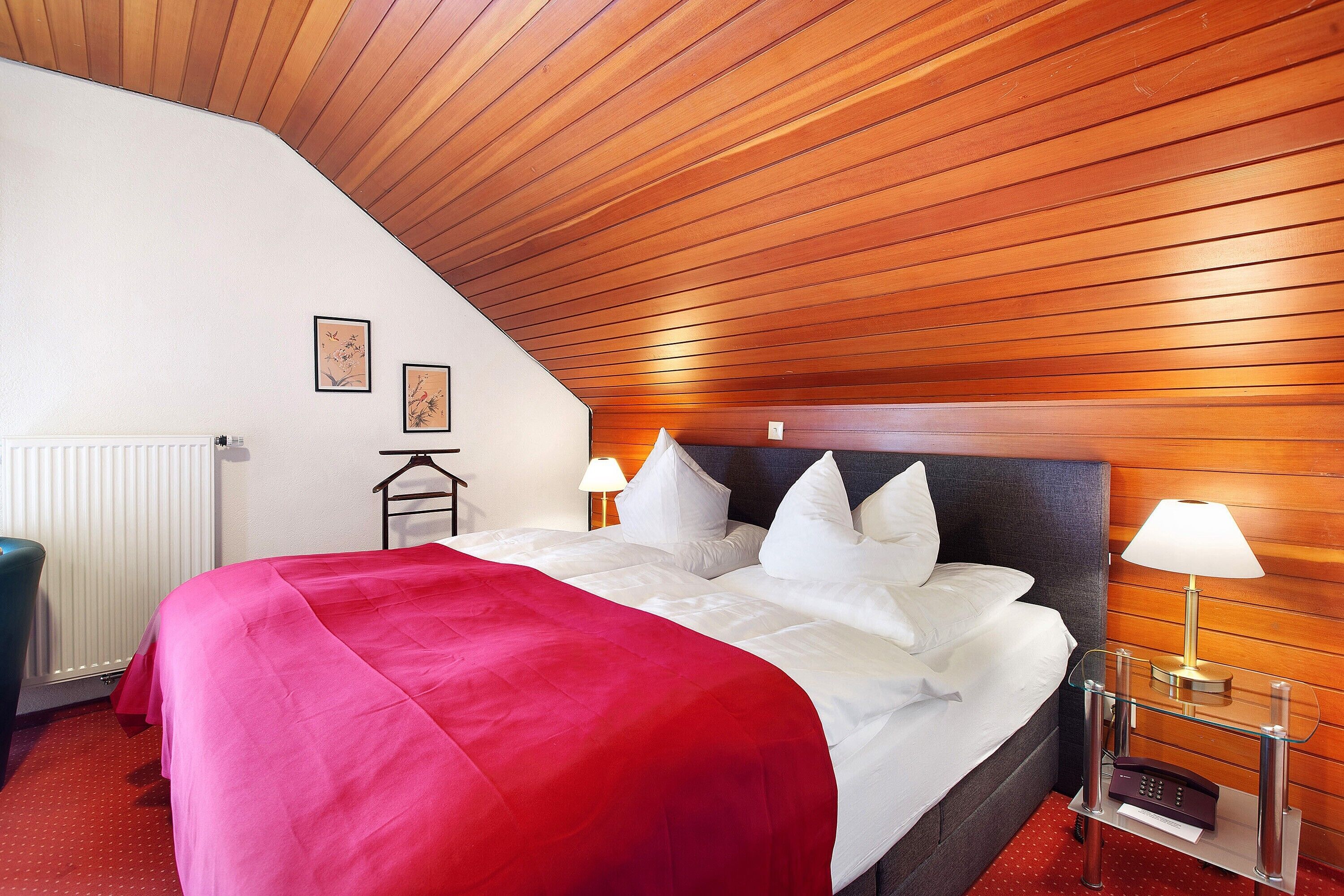 Une chambre d’hôtel avec un plafond en pente, un lit avec une literie blanche et un plaid rouge, une table de chevet avec une lampe et un radiateur.