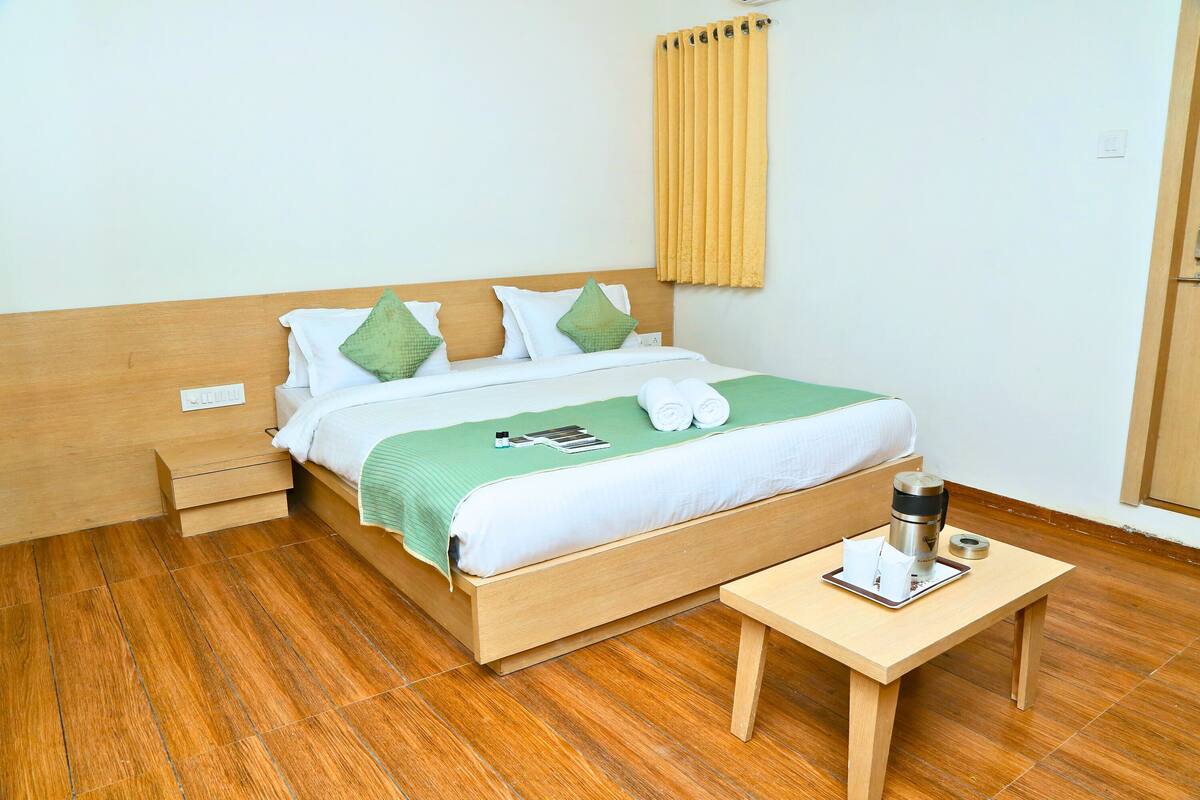 A neatly made bed with a green bedspread, white linens, and two green pillows. There is a wooden bedside table with a cup and a tray.