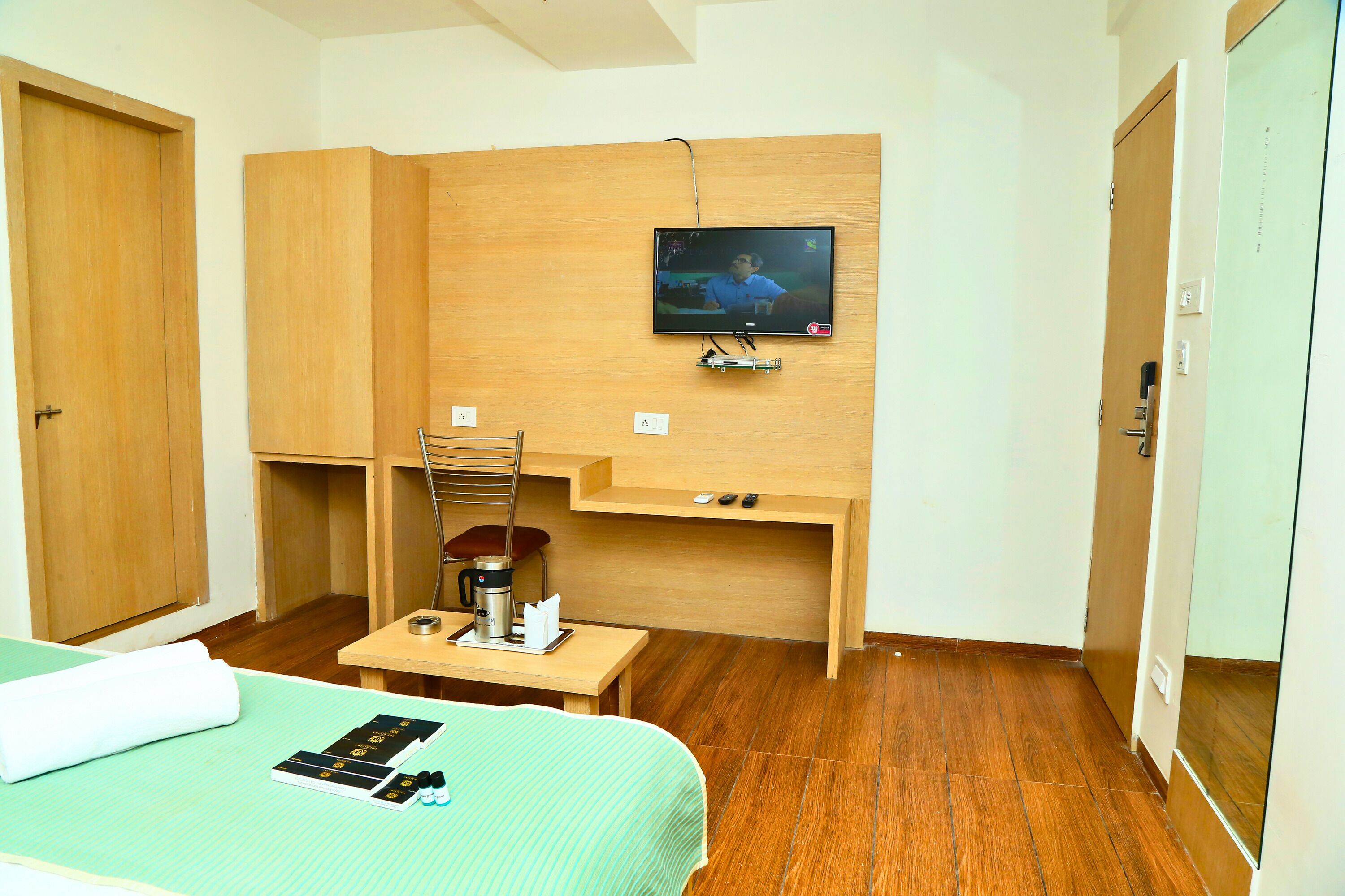 A compact hotel room with a bed, a desk with a chair, a television mounted on the wall, and a small table with a coffee maker.