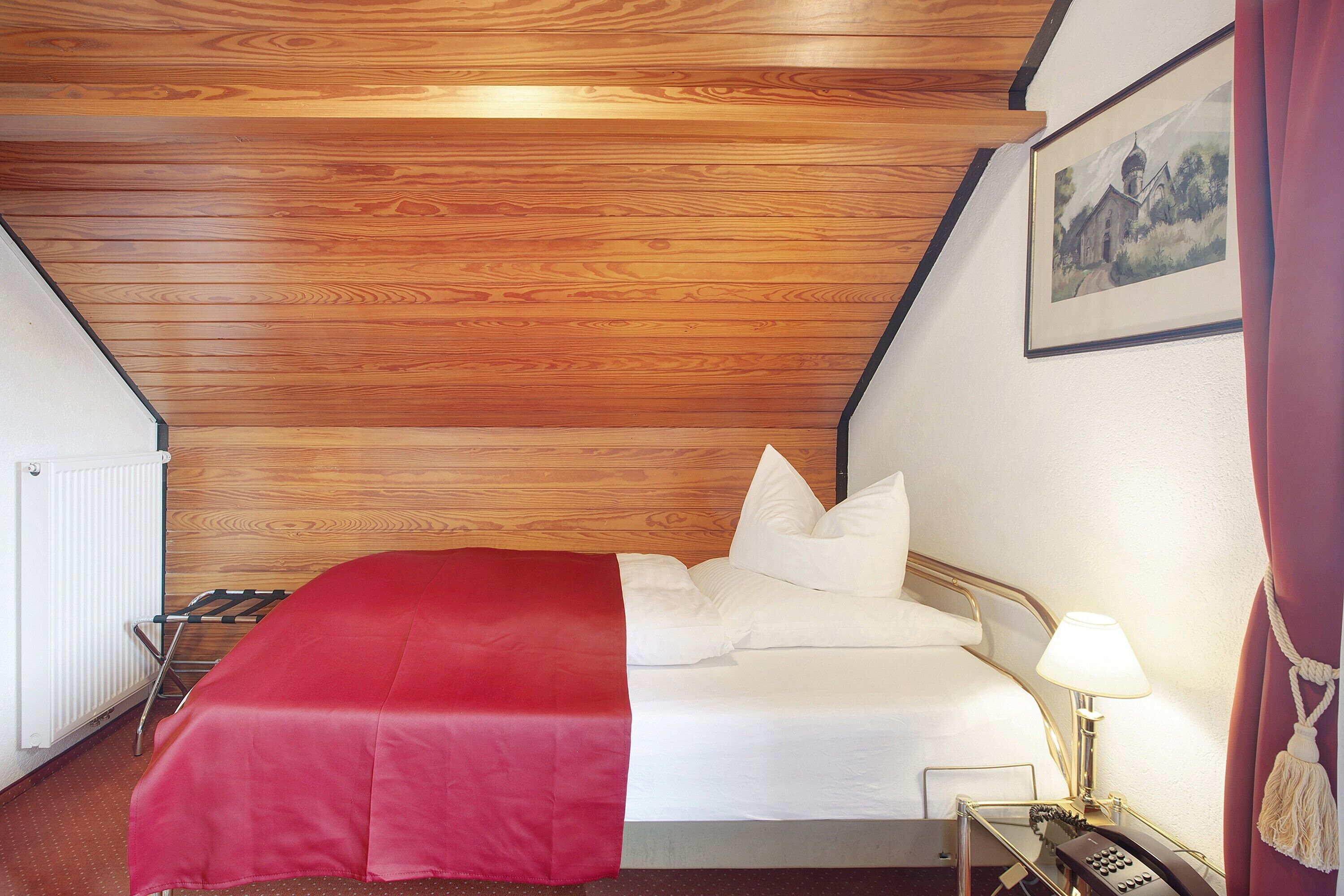 A hotel room with a wooden ceiling, a bed with white bedding, a red throw, a bedside lamp, and a painting on the wall.