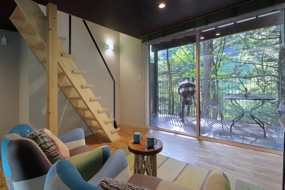 Una sala de estar moderna con una escalera de madera, un amplio ventanal corredero que da a una terraza exterior y un área de descanso con una pequeña mesa redonda.