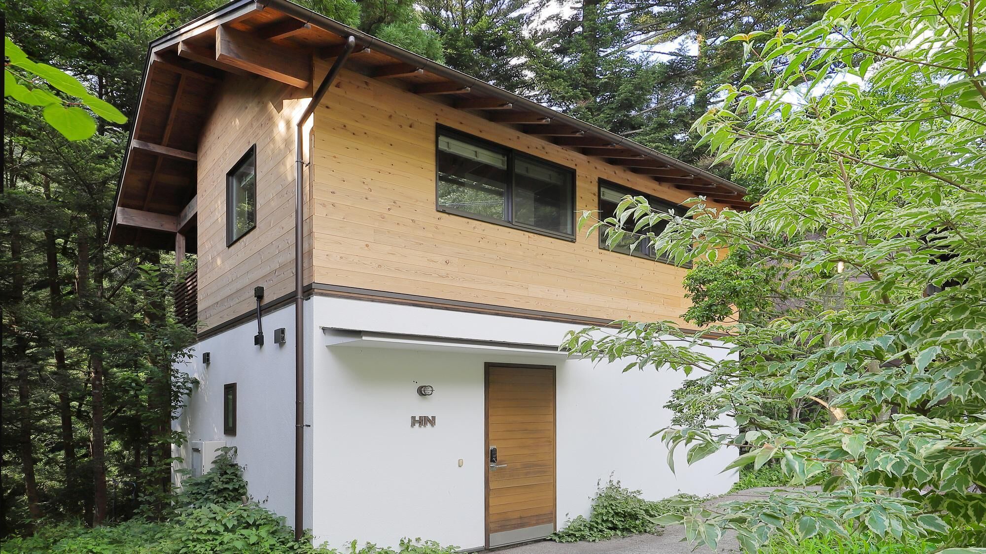 A two-story wooden house with a white lower level and a wooden upper level, surrounded by lush greenery.