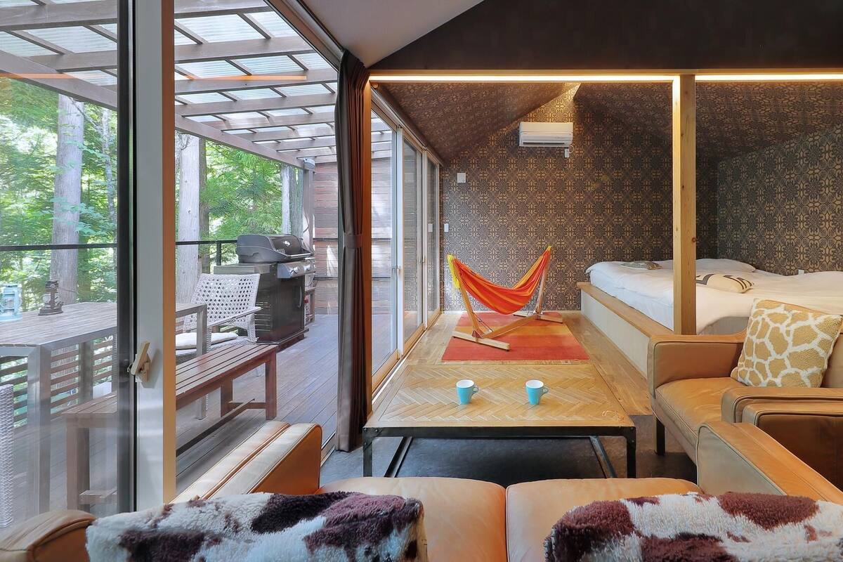 Una habitación moderna con puerta de vidrio que da a una terraza exterior, una hamaca, una cama, un sofá y una mesa con dos tazas.