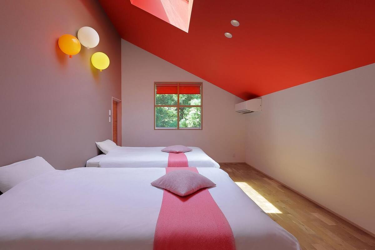 Una habitación con dos camas, una ventana que da a árboles y un techo rojo.