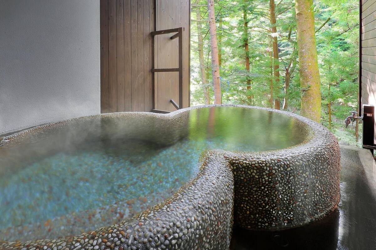 Un baño de aguas termales con revestimiento de guijarros, rodeado por una estructura de madera y vistas a una zona boscosa.