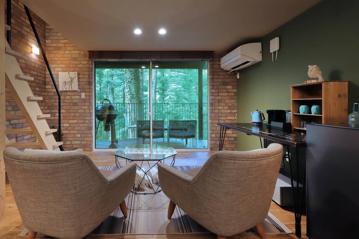 Una sala de estar moderna con una mesa de vidrio, dos sillones, una escalera con escalones de madera y vistas a un bosque a través de puertas de cristal.