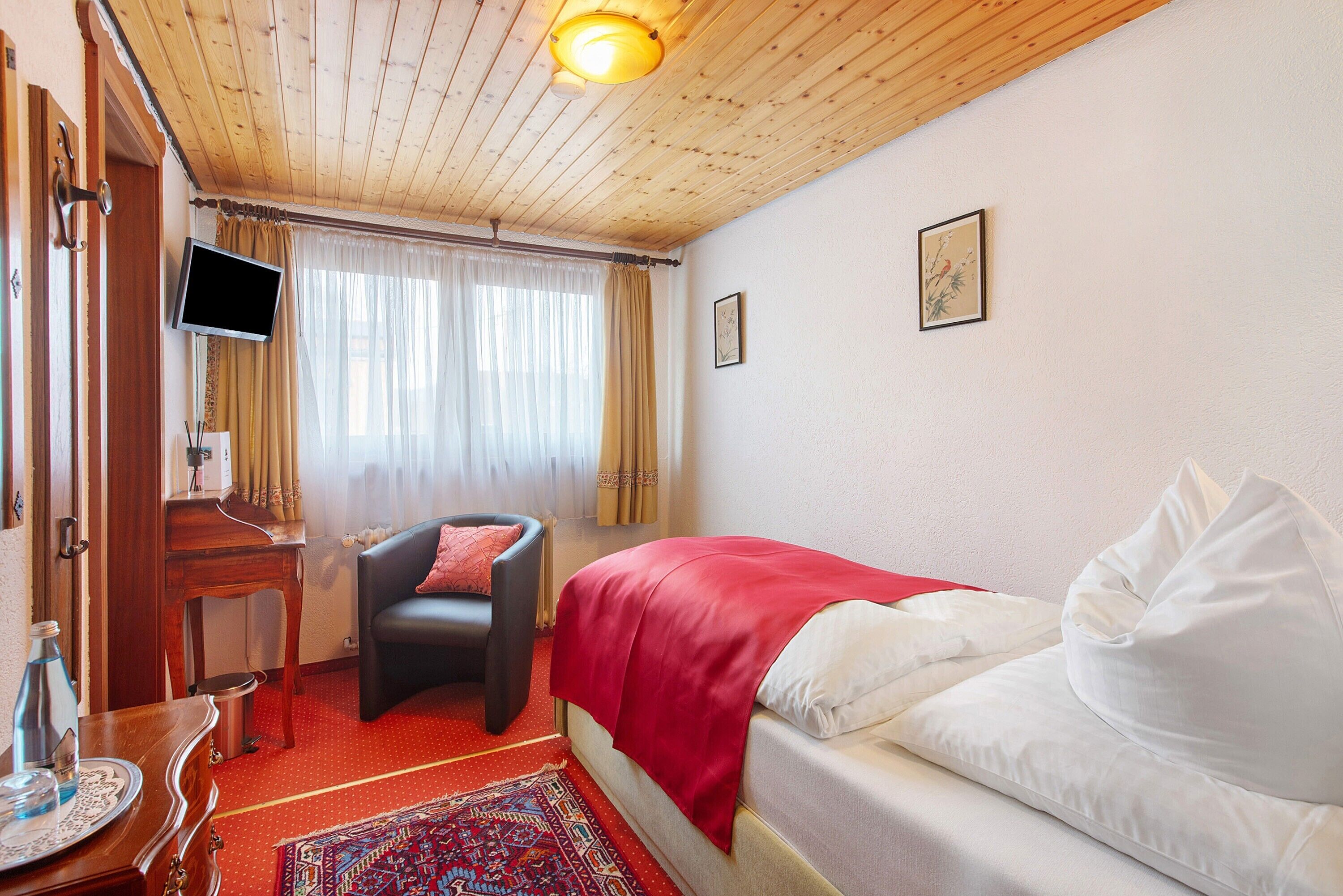 Habitación de hotel con una cama, una silla, un escritorio con televisor de pantalla plana, una ventana con cortinas y techo de madera.