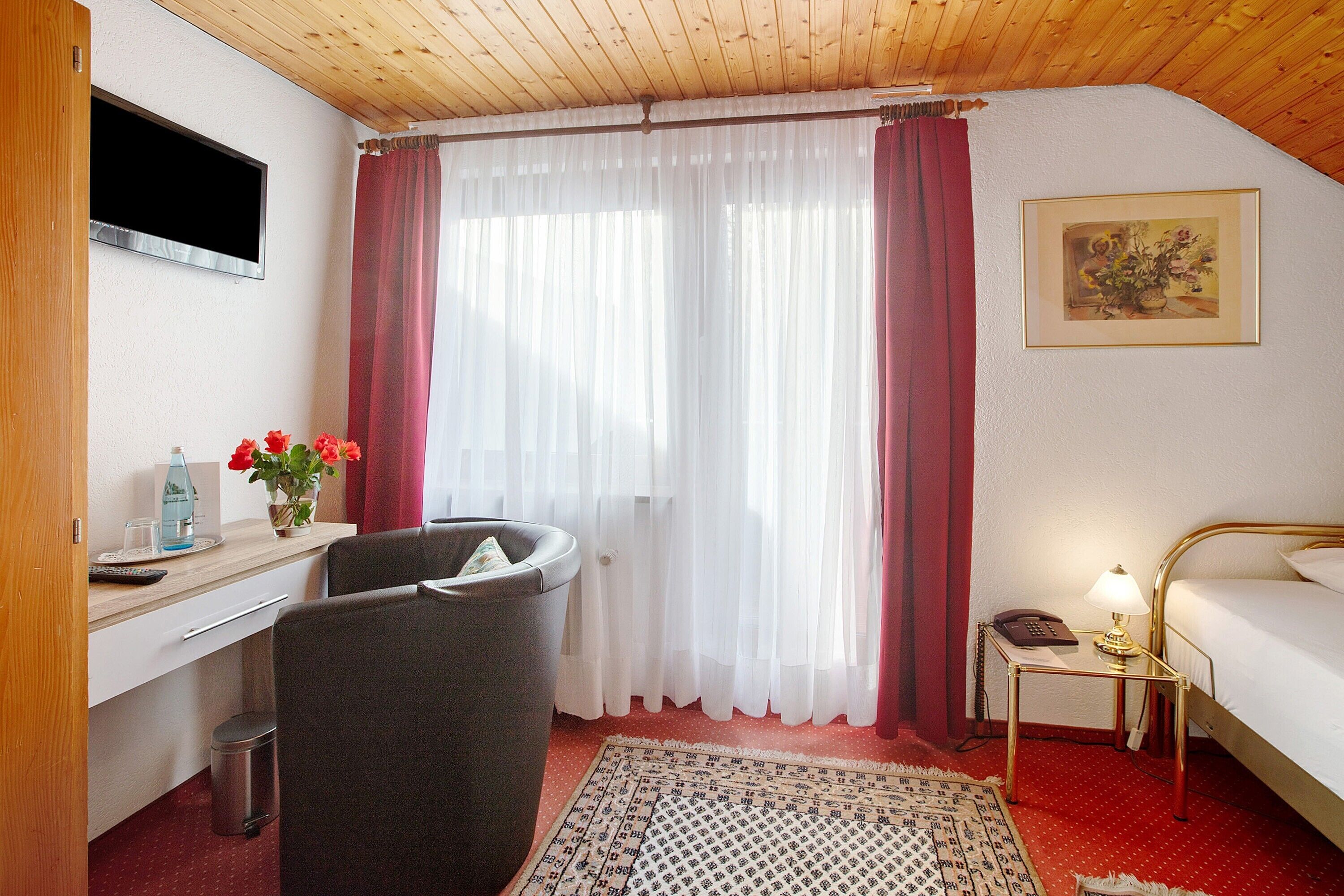 Une chambre d’hôtel comprenant un lit, un bureau avec une chaise, une poubelle, une petite table avec une lampe, un tableau au mur et une fenêtre avec des rideaux.