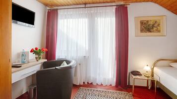 Une chambre d’hôtel comprenant un lit, un bureau avec une chaise, une poubelle, une petite table avec une lampe, un tableau au mur et une fenêtre avec des rideaux.