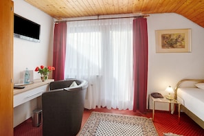 Una camera d'albergo con un letto, una scrivania con una sedia, un cestino, un tavolino con una lampada, un quadro appeso al muro e una finestra con tende.