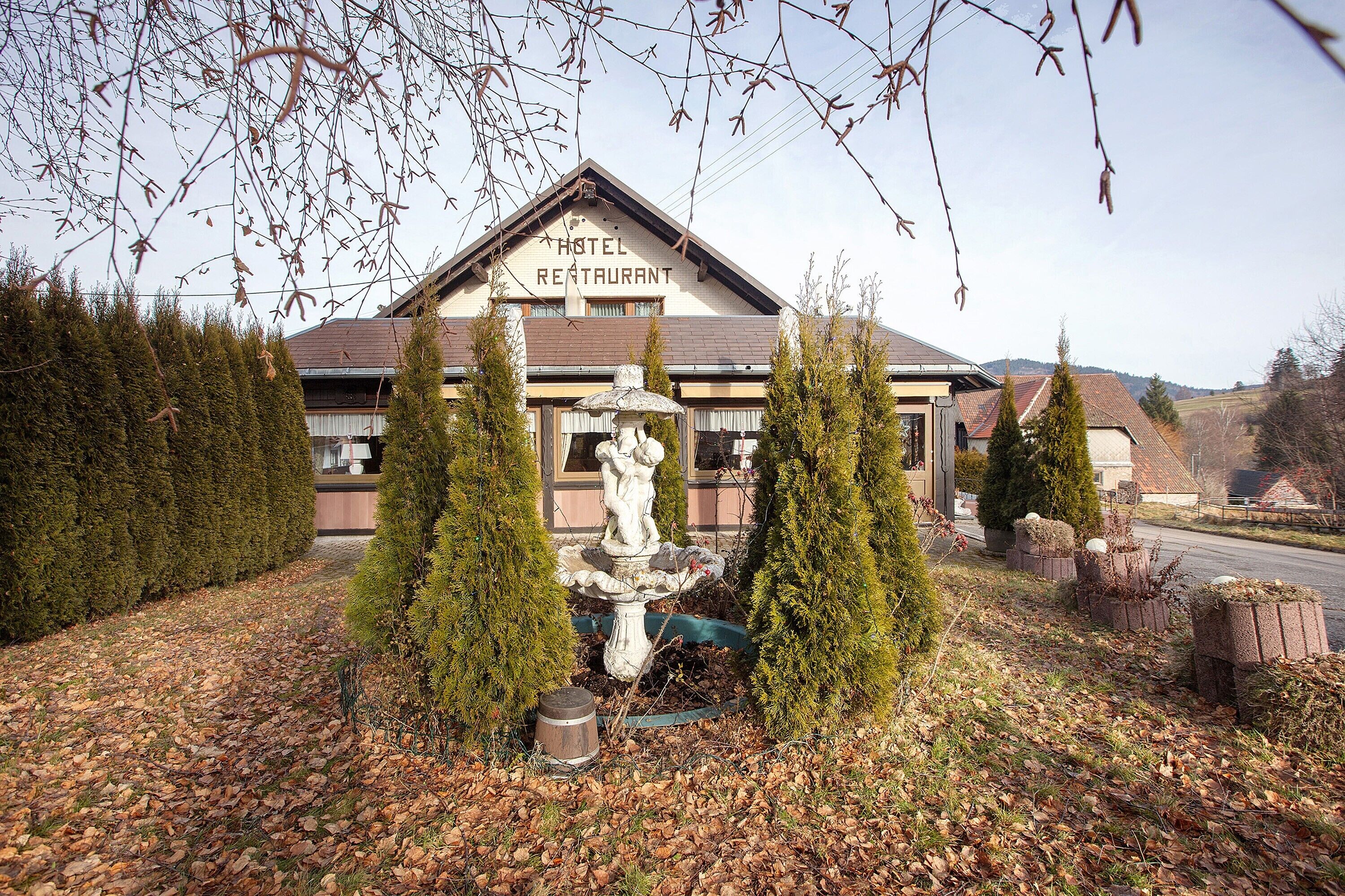 Un hôtel avec une enseigne indiquant « HÔTEL-RESTAURANT », entouré de haies soigneusement taillées et d’une fontaine en pierre.