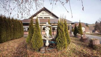 Un hôtel avec une enseigne indiquant « HÔTEL-RESTAURANT », entouré de haies soigneusement taillées et d’une fontaine en pierre.