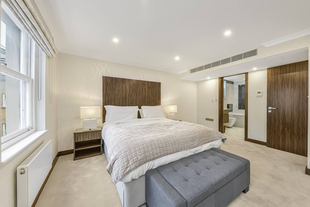 Ein modernes Schlafzimmer mit einem großen Bett, Nachttischen, einer Bank, einem Kleiderschrank und einem durch eine offene Tür erreichbaren Badezimmer.
