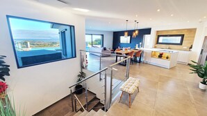 A modern kitchen with a central island, a dining area with a sofa, and a staircase leading to an upper level with a balcony. - Villa de prestige en Haute-Corse - Sea view & exceptional amenities (Penta-di-Casinca)