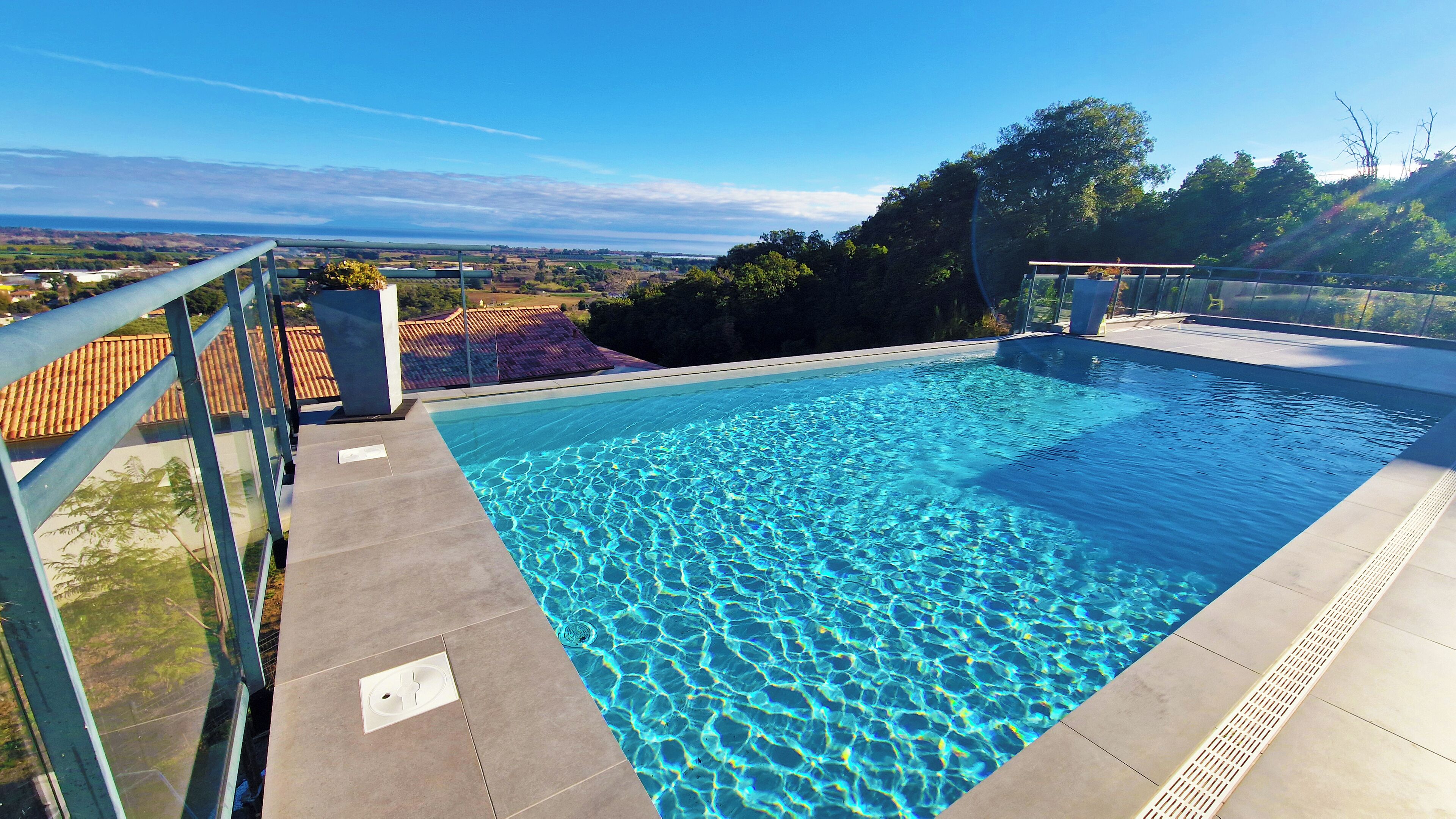 Ein Schwimmbecken mit klarem, blauem Wasser, umgeben von einer Glasbrüstung, mit Blick auf eine malerische Landschaft aus Gebäuden und Bäumen.