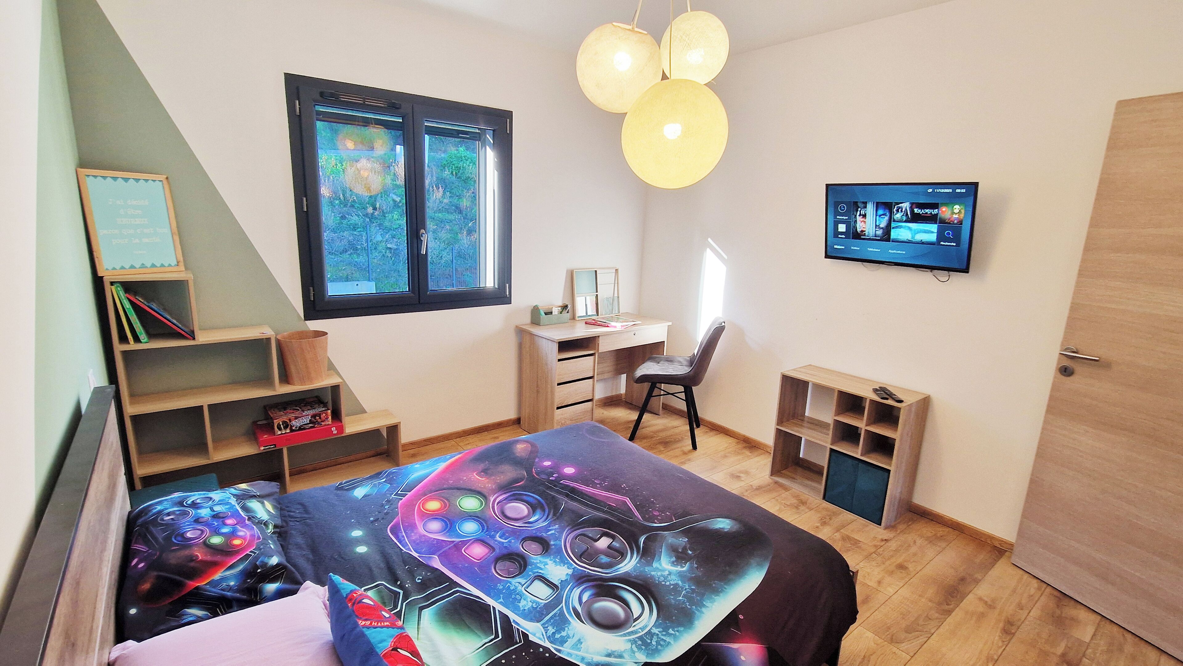 Une chambre moderne équipée d’un lit conçu pour les jeux vidéo, d’un bureau, d’une chaise, d’un téléviseur et d’une fenêtre donnant sur un paysage verdoyant.