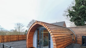 Uma cabana de madeira com entrada arqueada e porta de vidro, cercada por um portão de metal e um pátio pavimentado.