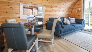 Interior acolhedor de cabana com teto de madeira, mesa de jantar com cadeiras, sofá azul e janela com vista para um jardim.