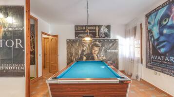 Una mesa de billar en una habitación con suelo de madera, carteles en las paredes y una puerta que da a otra estancia.