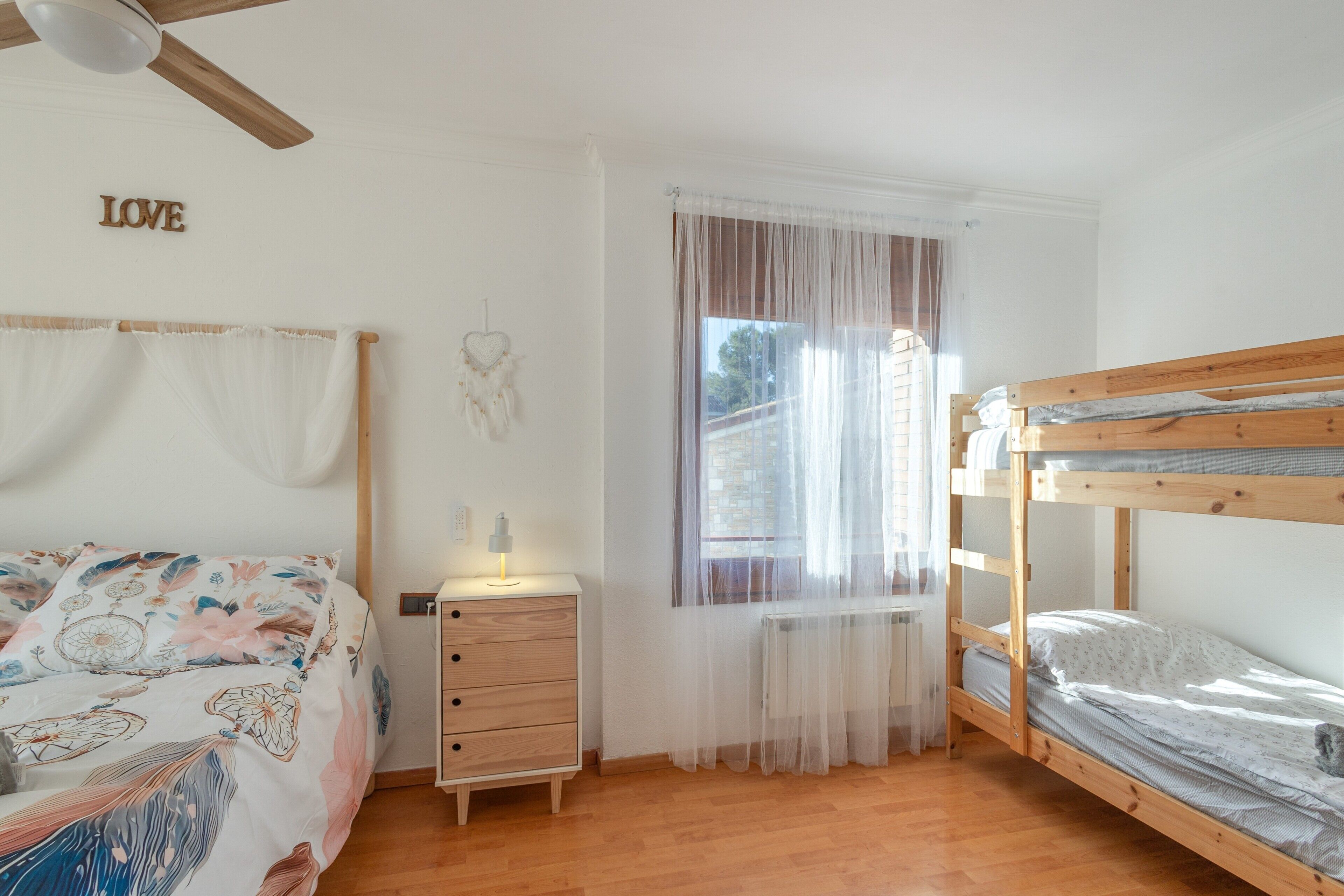 Un dormitorio con una cama individual superpuesta, una mesita de noche con lámpara, un aparador, una ventana con cortinas traslúcidas y un atrapasueños colgado en la pared.
