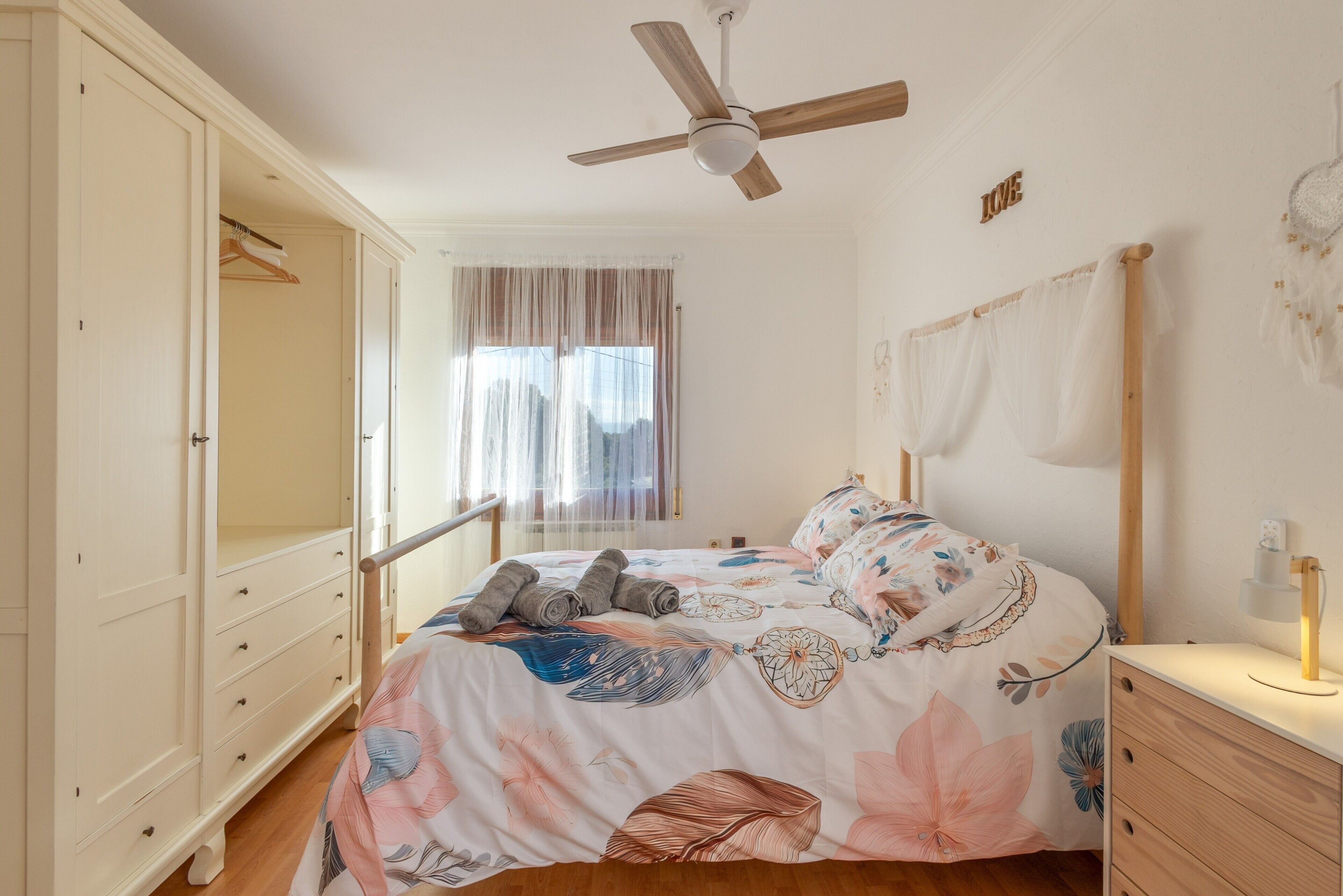 Een slaapkamer met een bed, een kledingkast, een plafondventilator, een nachtkastje en een raam met gordijnen.