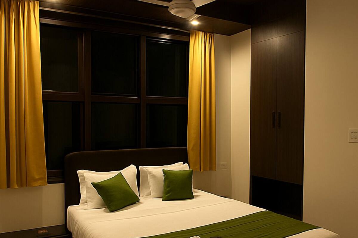 A neatly made bed with a green throw, a ceiling fan, and a window with yellow curtains.