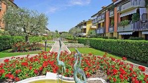 A pathway leading through a garden with red flowers and a fountain with two statues. - Villaggio dei Fiori - Viola (Sirmione)
