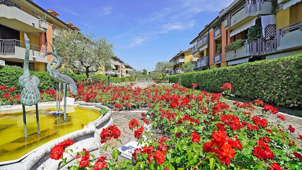 A courtyard with red flowers, a fountain, and apartment buildings. - Villaggio dei Fiori - Viola (Sirmione)