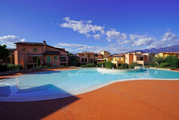 A swimming pool with a curved design, surrounded by terracotta-tiled walkways and buildings with Mediterranean-style architecture. - Borgo del Torchio in Manerba del Garda (Manerba del Garda)