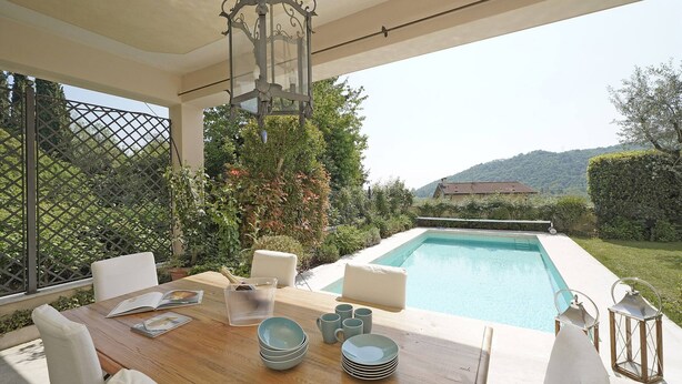 A poolside dining area with a wooden table, white cushions, and a view of a swimming pool and greenery. - Villa San Carlo - Gardagate (Roe Volciano)