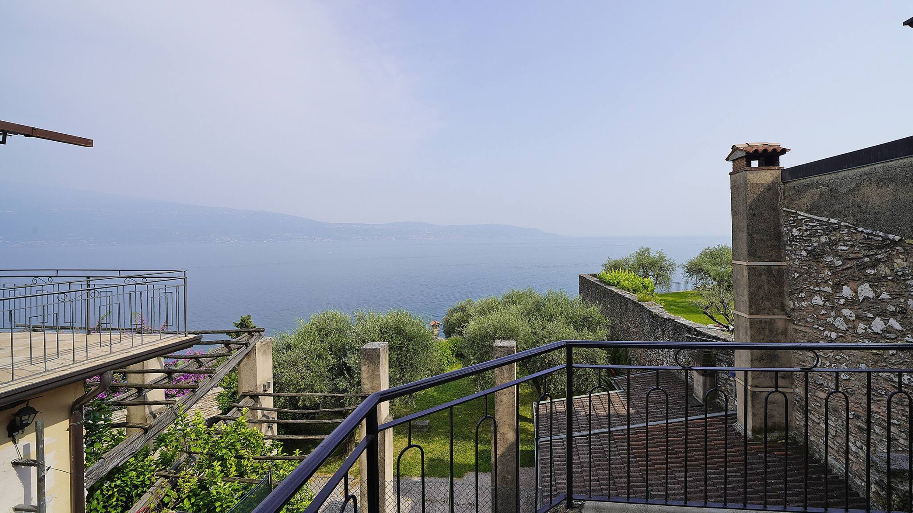 A terrace with a stone wall and a metal railing overlooking a body of water and distant mountains.