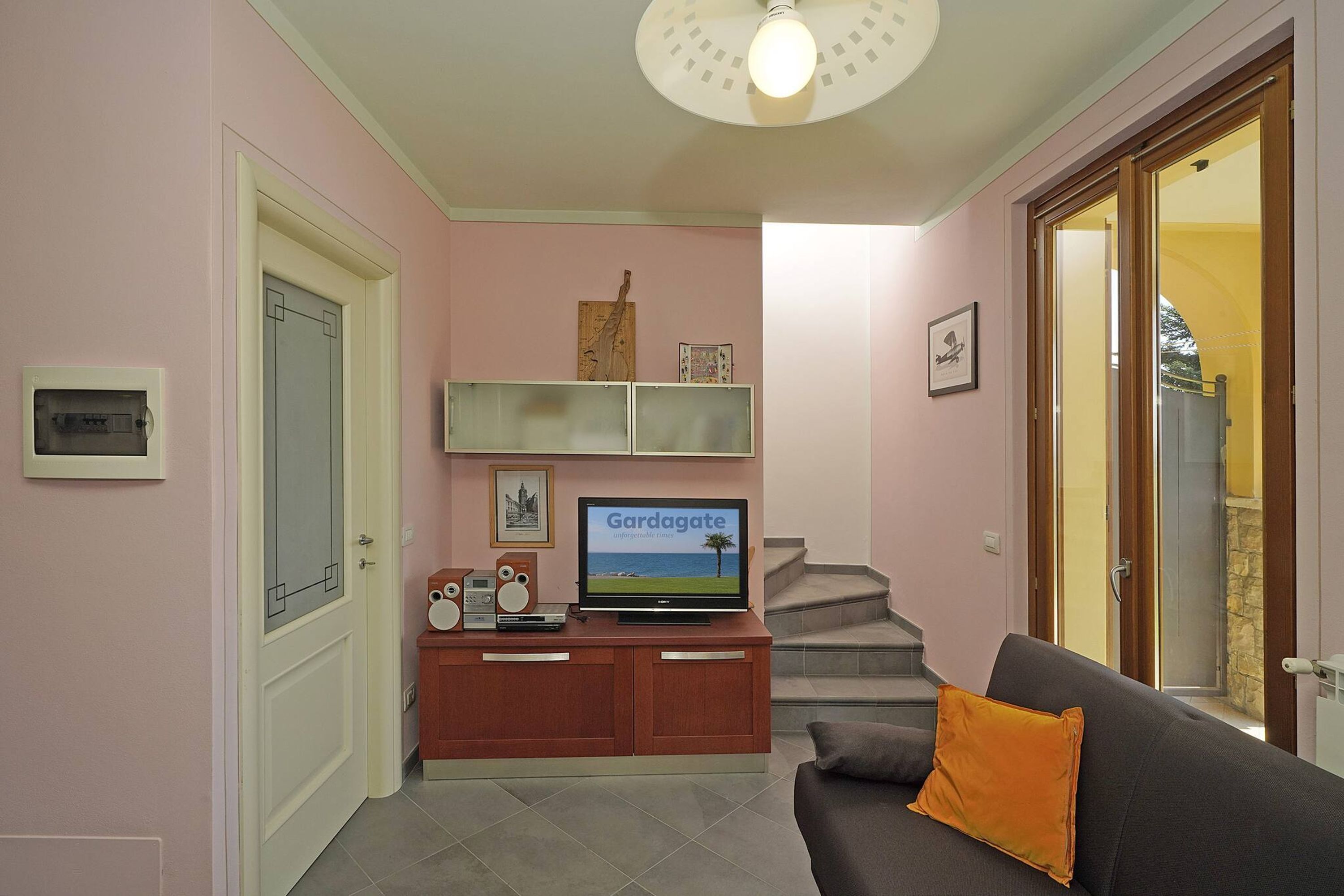 A living room with a sofa, a television, a cabinet, and a staircase leading to an outdoor area.