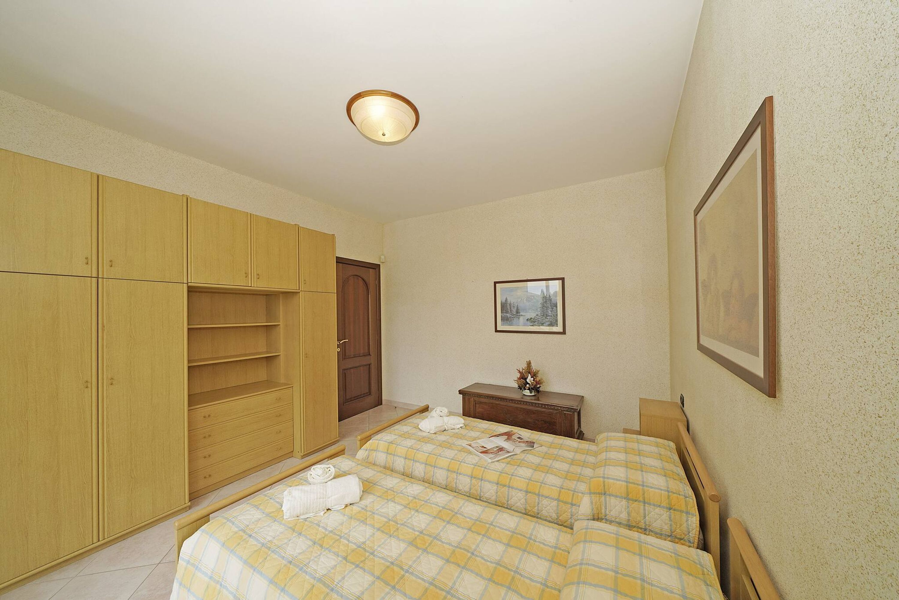 Un dormitorio con dos camas, un armario, una mesita con un florero y un cuadro en la pared.