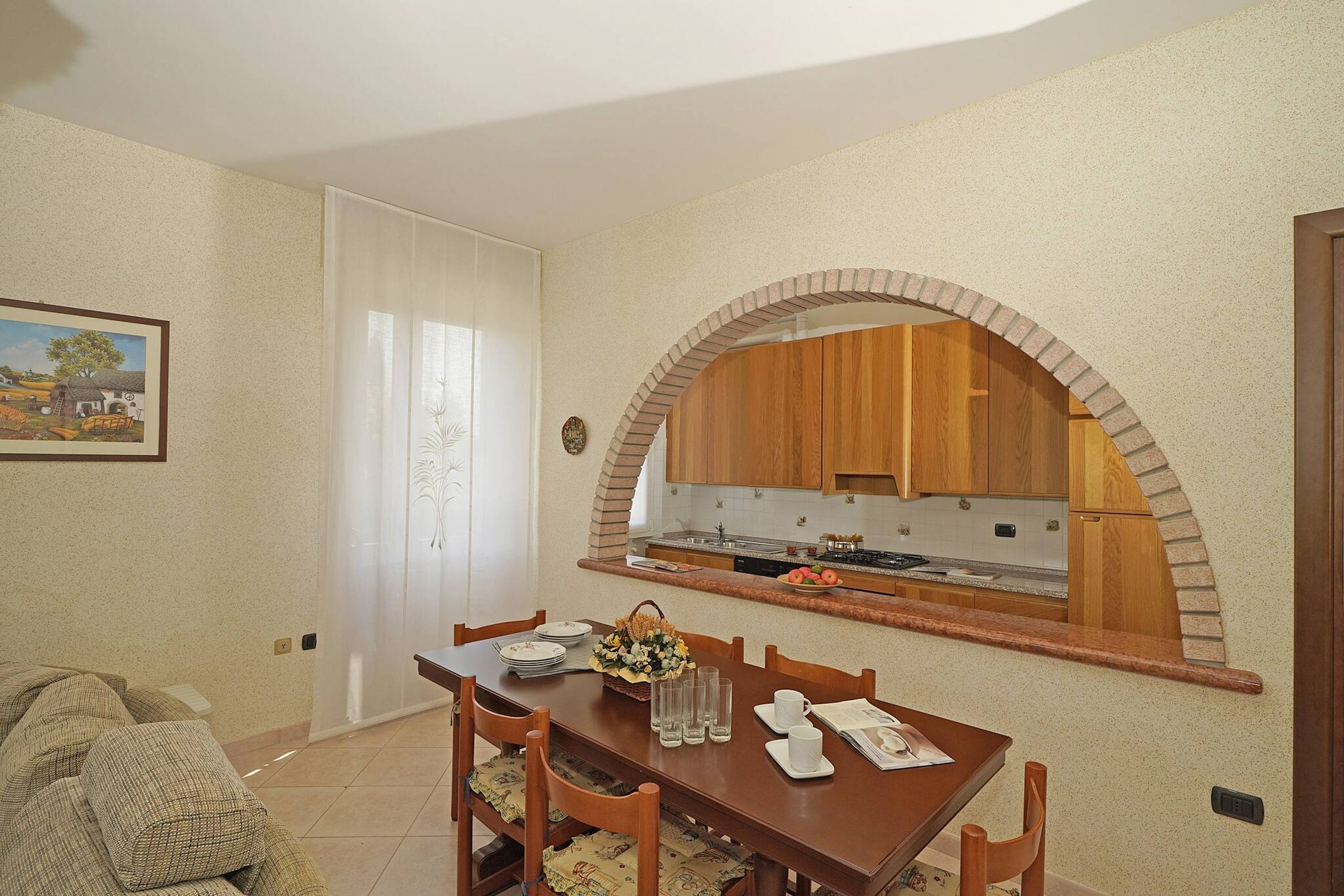 Ein Essbereich mit Holztisch, Stühlen und einer durch ein Rundbogenfenster einsehbaren Küche.