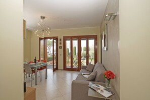 Una sala de estar moderna con sofá, mesa de comedor y una araña. Se ve un balcón con mesa y sillas a través de puertas de cristal.