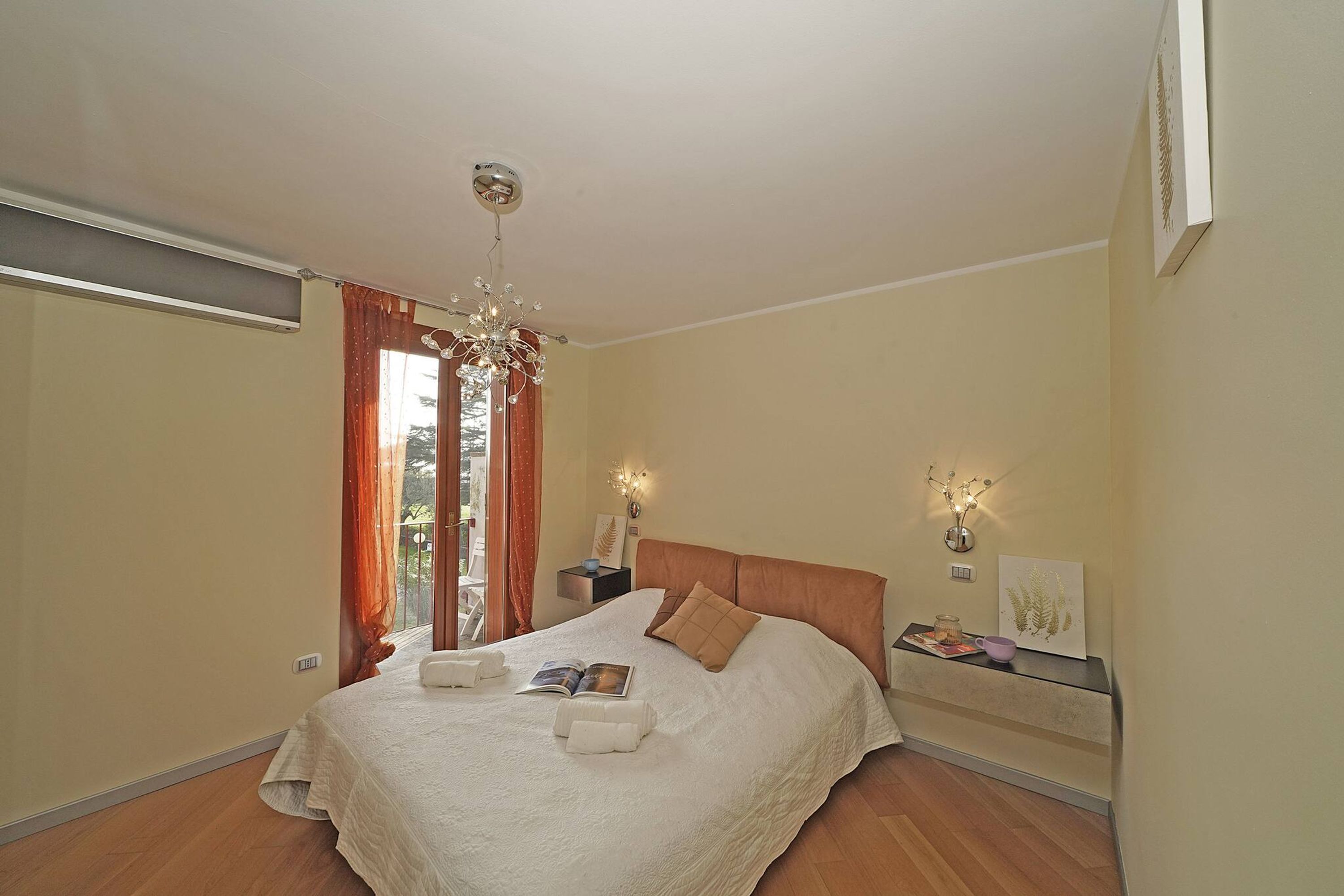 Un dormitorio ordenado con una cama grande, una mesita de noche con lámpara y vistas al exterior por una ventana.
